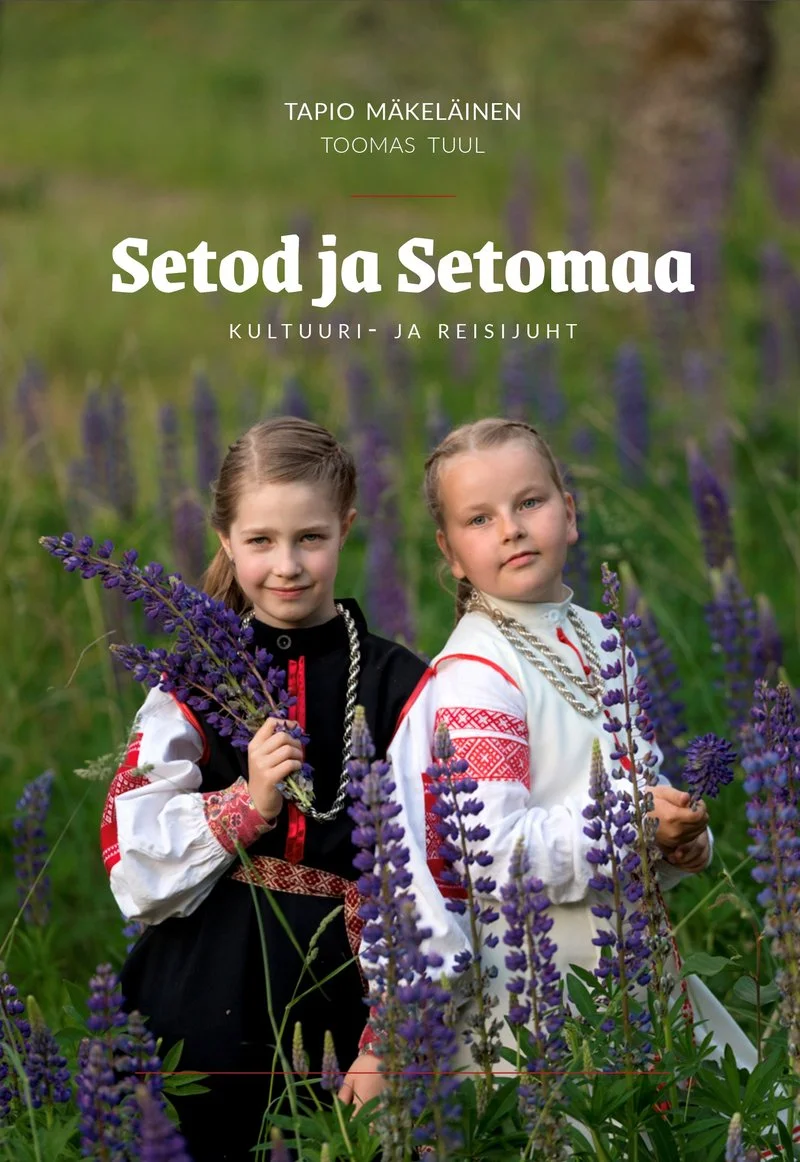 Setod ja Setomaa. Kultuuri- ja reisijuht - Tapio Mäkeläinen, Toomas Tuul (Postimehe kirjastus, 2026). Kaks tüdrukut seto rahvariieteis seisavad õitsvate lillede keskel rohelisel niidunurmel. Foto on soe ja looduslähedane, rõhutades seto kultuuripärandi autentsust.