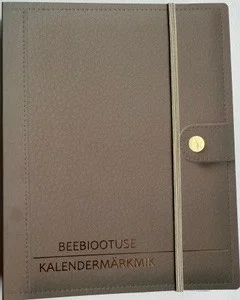 Beebiootuse kalendermärkmik - Brigitta Leif (LEIF KIRJASTUS, 2026). Pruunikas-halli kunstnahast märkmiku kaas kuldse tekstiga, millel on küljes eraldi pliiatsihoidja tasku ja kuldne nuppsulg.